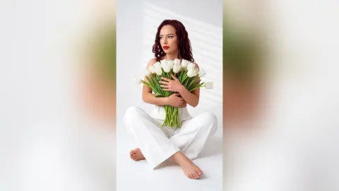 Elegant woman in white outfit holding a large bouquet of white tulips in a bright, minimalist studio setting.