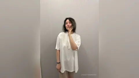 Young woman in an oversized white t-shirt posing playfully against a minimalist grey studio background.