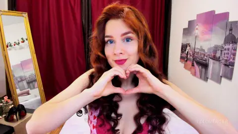 Smiling redhead woman in a pink top making a heart gesture against red curtains in a cozy room.