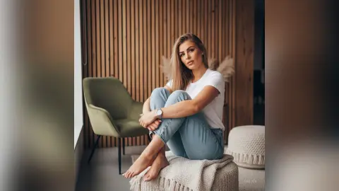 Hailey poses sitting on a white textured stool wearing a white t-shirt and blue jeans, with her bare feet visible against a wooden slatted wall.