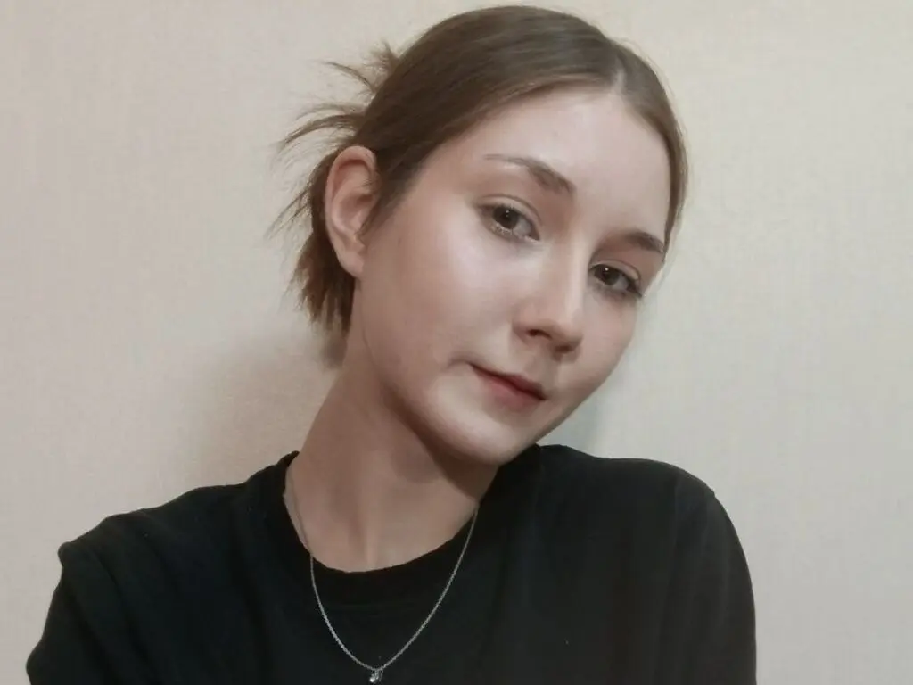 Young woman with brown hair in a black t-shirt posing against a neutral wall with a soft, natural look.
