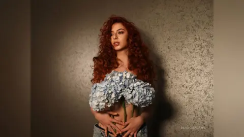 Redheaded woman in denim holding a large bouquet of blue hydrangeas against a textured wall in soft lighting.