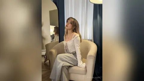 Young woman in a white lace blouse and grey trousers sitting in a cozy, sunlit room with a dreamy mood.
