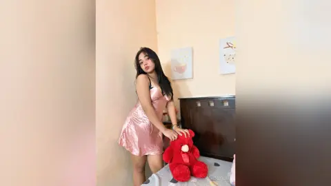Young woman in a pink satin slip dress sitting on a bed with a red teddy bear in a cozy bedroom setting.