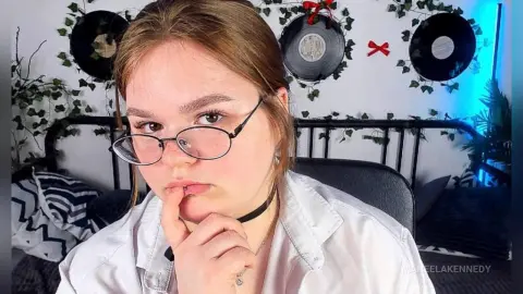 ManeelaKennedy wears glasses and a black choker while looking at the camera in her bedroom.