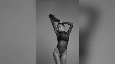 MilanaVolkova poses in a black sheer bodysuit with her arms raised behind her head against a plain grey background.