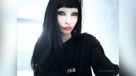 Goth girl with long black hair and nose ring wearing a black t-shirt in a moody, minimalist setting.