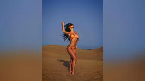 Slim woman in an orange bikini and sunglasses posing confidently in a sunny desert sand dune landscape.