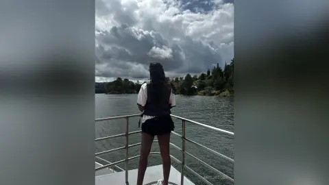 Woman in white top and black shorts standing on a boat deck overlooking a lake under a moody cloudy sky.