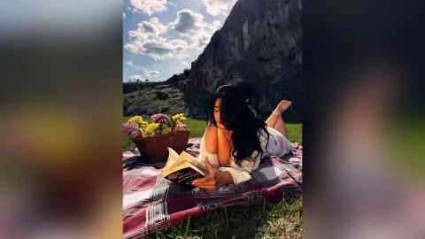 Relaxed woman in a white dress reading a book on a picnic blanket in a sunny meadow near a mountain.