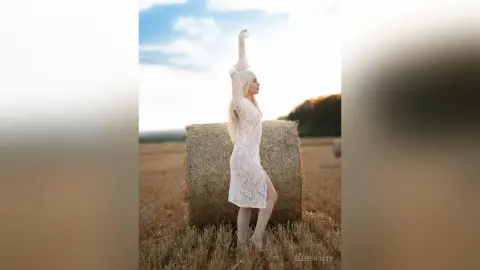 Blonde woman in a white lace dress posing gracefully by a hay bale in a golden sunlit field.