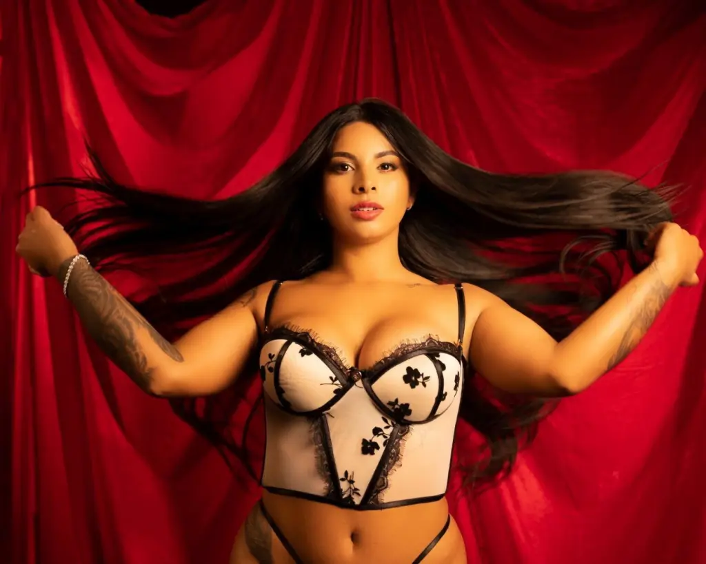 Gaia_Grey poses with long black hair, wearing a black lace bra and a black choker, showing off her cleavage against a warm red background.
