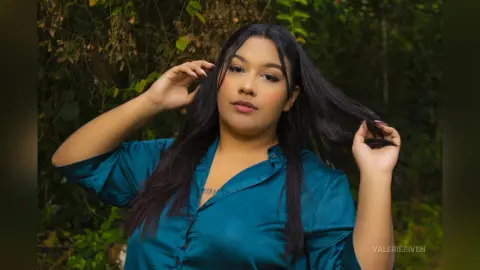Young woman with long dark hair wearing a teal button-down shirt in a lush green forest setting.