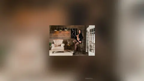 Elegant woman in a black blazer sitting on a barstool in a modern rustic living room with wood walls.