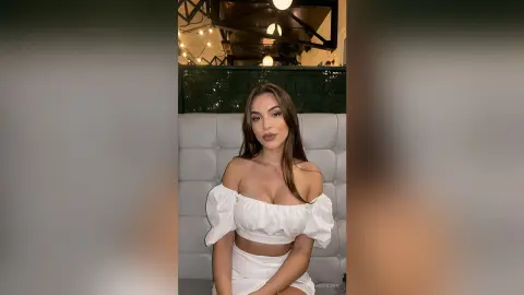 Elegant woman in a white off-the-shoulder crop top sitting in a chic, modern restaurant setting.