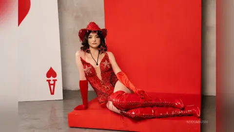 Woman in a sparkling red sequin bodysuit and cowboy hat sitting against a red heart backdrop.