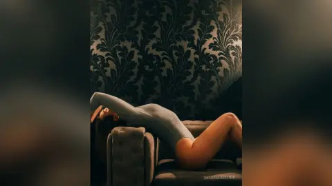 Woman in a grey bodysuit relaxing on a sofa against dark floral wallpaper in a moody, intimate setting.