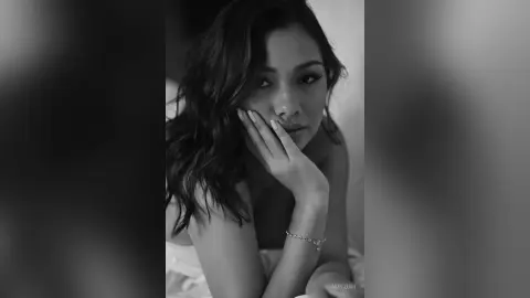 Moody black and white portrait of a woman with wavy hair resting her chin on her hand in a soft indoor setting.