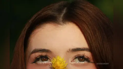 Close-up portrait of a woman with brown hair and long eyelashes holding a yellow flower near her face.