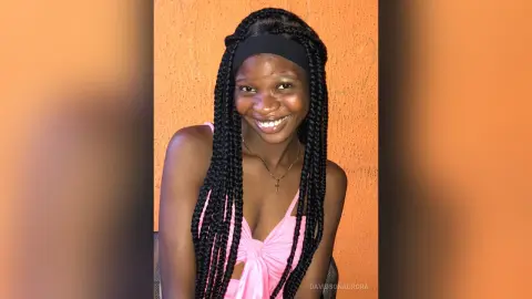 Smiling young Black woman with long braids wearing a pink tank top against an orange wall.