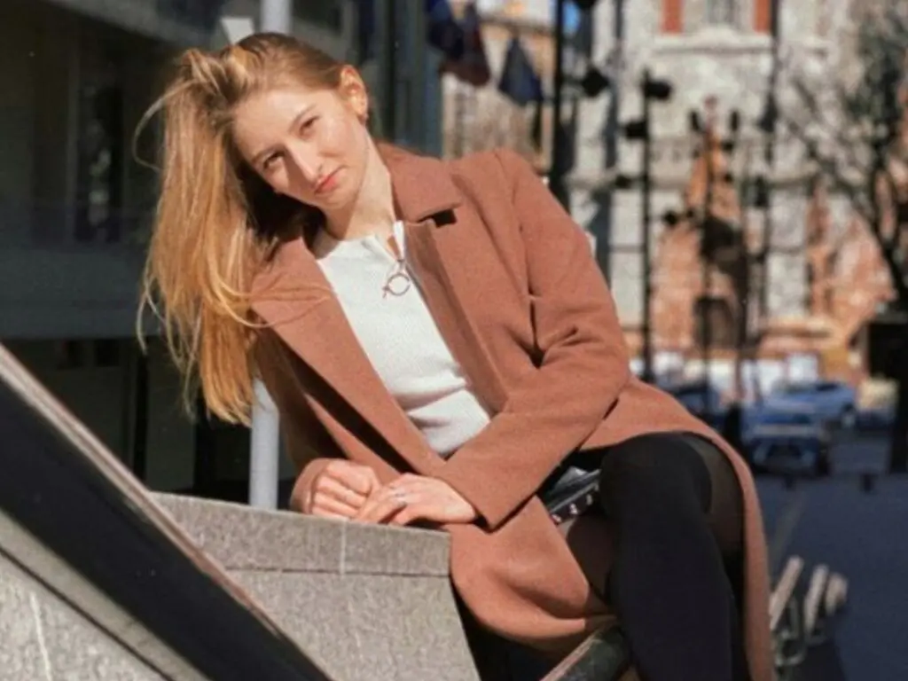 KaterineKoen poses outdoors in a long brown coat and white top, letting her long blonde hair hang down as she leans against a stone ledge.