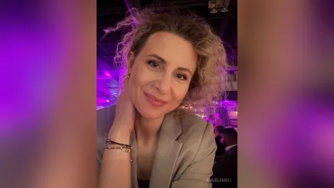 AngelEmeli — AngelEmeli smiles at the camera with her hand on her neck, wearing a grey blazer in a room with purple lighting.