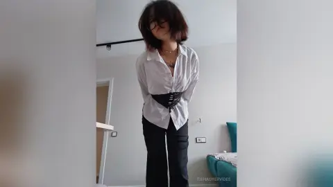 TishaOyervides wears a white button-down shirt and black pants with a black corset, standing in a bright room with dark hair.