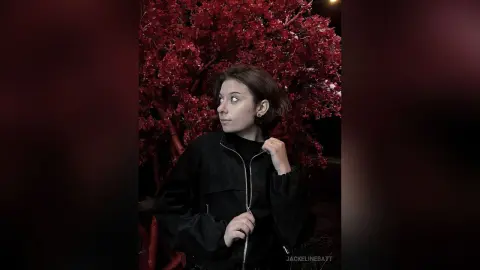 Young woman in a black jacket looking away against a backdrop of vibrant red autumn leaves in a moody setting.