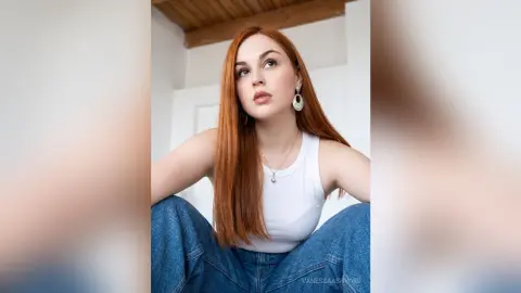 Redheaded woman in a white tank top and blue jeans looking thoughtfully in a bright, minimalist room.