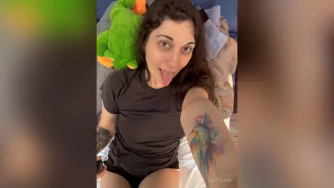 Smiling woman in a black t-shirt relaxing on a bed with a green plush toy in a cozy indoor setting.