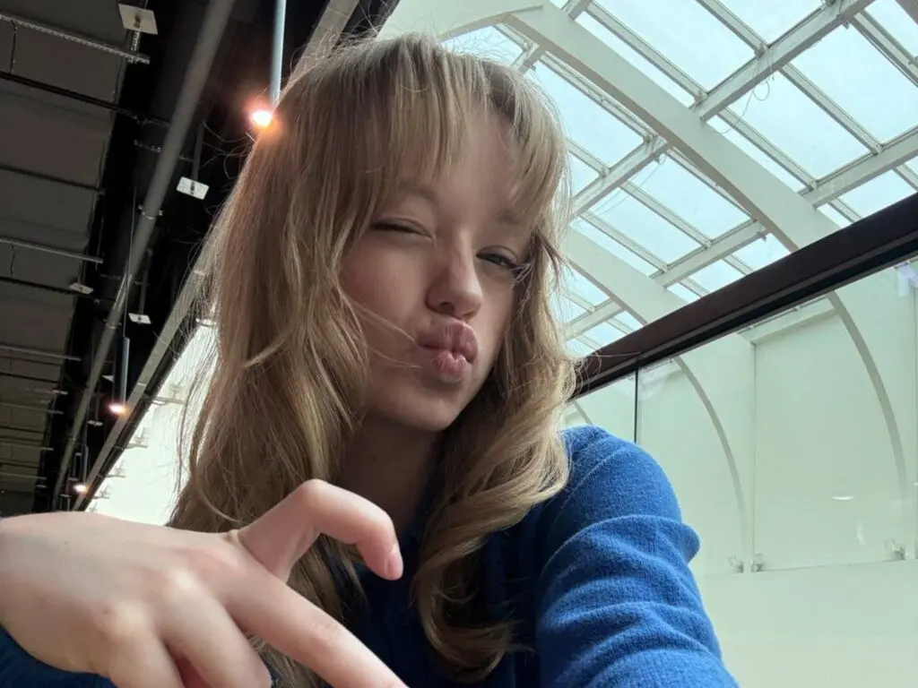 Blonde woman in a blue sweater making a playful kissy face in a bright, modern glass atrium.
