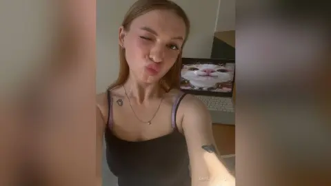 DahliaSackey pouts her lips and winks at the camera, wearing a black tank top and a silver necklace in a room with a laptop visible behind her.