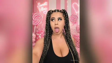 Playful woman with long blonde braids sticking her tongue out in a pink candy-themed setting.