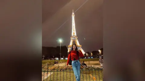 LucyMartin stands in front of the lit Eiffel Tower at night wearing a red long-sleeve top and blue jeans.