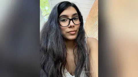 Young woman with long dark hair and black glasses wearing a white top in a bright outdoor setting.