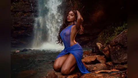 Elegant woman in a flowing blue gown posing gracefully by a misty waterfall in a lush, moody forest setting.
