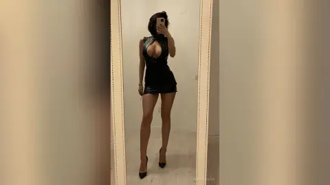 Woman in a sleek black leather mini dress posing for a mirror selfie in a minimalist modern room.