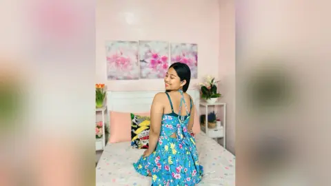 Smiling woman in a blue floral summer dress posing in a bright, cozy bedroom.