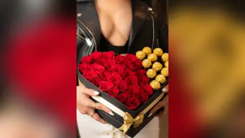 RebecaVillalobos holds a black box filled with red roses against her cleavage while wearing a black leather jacket.