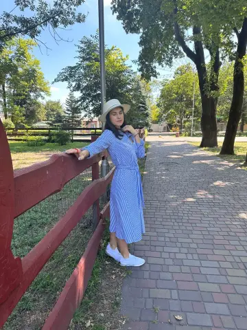 Woman in a blue striped summer dress and white hat leaning on a red fence in a sunny park setting.