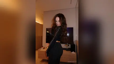 Stylish woman in a black long-sleeve top and pinstripe trousers posing in a modern, dimly lit living room.