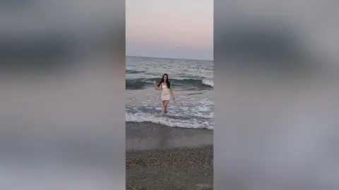 SaraTray stands in the shallow ocean waves at a beach, wearing a short white sundress with her long dark hair down.