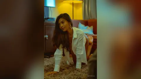 Moody portrait of a woman in a white button-down shirt sitting on a rug in a warm, dimly lit room.