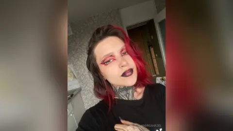 AndreaHazel poses with red hair and dark lipstick, wearing a black t-shirt and showing her neck tattoos.