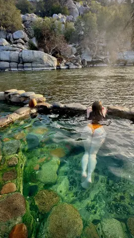 Woman in orange bikini relaxing in a clear mountain spring surrounded by natural rocks and lush greenery.