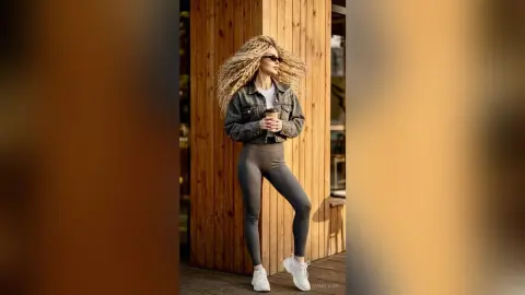 Stylish woman in denim jacket and grey leggings holding coffee against a rustic wooden wall background.