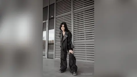 Edgy woman in black leather jacket and baggy trousers posing against a modern slatted metal wall background.