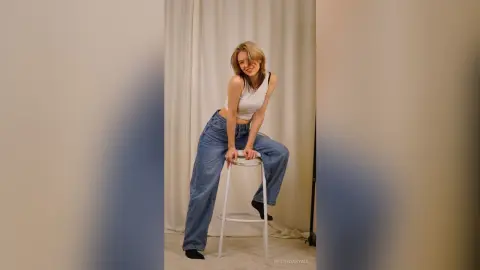 Smiling young woman in white tank top and baggy blue jeans posing on a white stool in a bright studio.