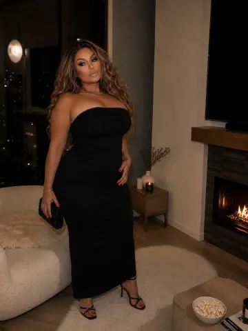 Samistaxxx poses in a tight black strapless dress and high heels in a living room near a fireplace.
