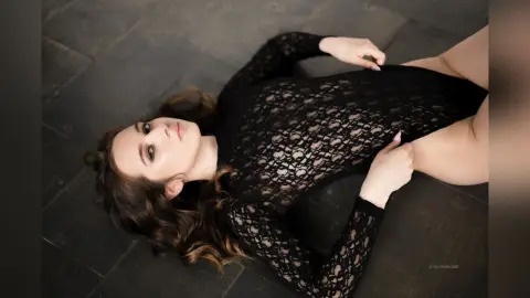 StacyHarland lies on a dark floor wearing a long-sleeved black lace bodysuit that shows her skin through the lace pattern.